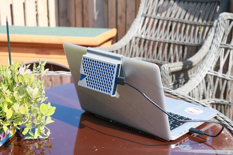 May include: A silver laptop computer with a blue and silver cooling fan attached to the back. The laptop is sitting on a wooden table with a green plant in a pot to the left. The laptop is plugged into an outlet with a black cord.