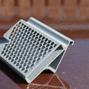 May include: A gray 3D printed phone stand with a honeycomb design. The stand is angled and has a raised lip on the top edge.