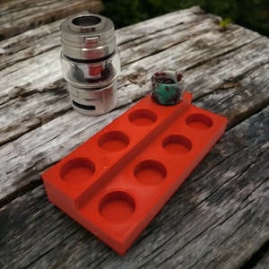 May include: Red plastic organizer with eight round compartments for storing vape tanks and coils. A silver vape tank and a green and brown drip tip are sitting on the organizer.