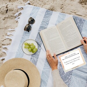 May include: A beach scene featuring a striped blanket, an open book, sunglasses, and a straw hat. A small plate of green grapes is placed nearby. A card with the text "BOOK CLUB QUESTION" is visible.