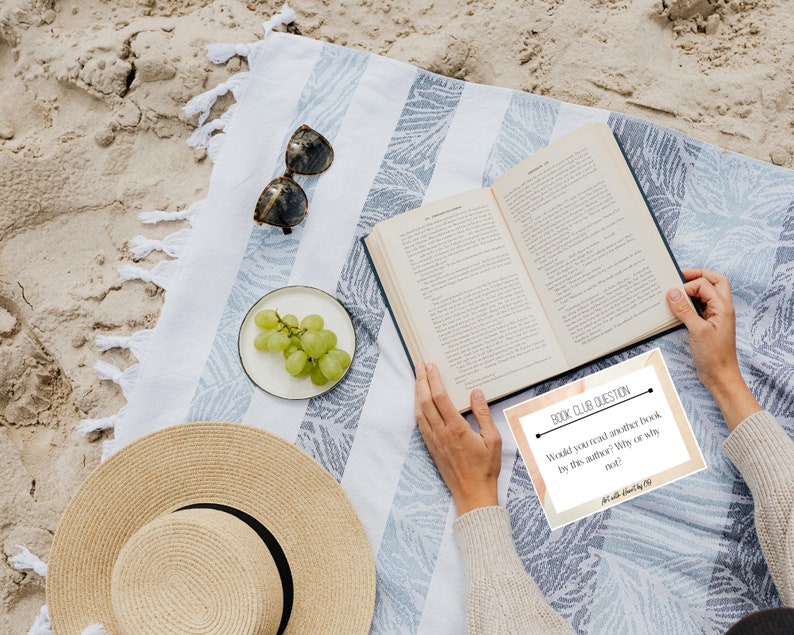 May include: A beach scene featuring a striped blanket, an open book, sunglasses, and a straw hat. A small plate of green grapes is placed nearby. A card with the text "BOOK CLUB QUESTION" is visible.