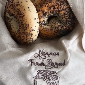 Könnte beinhalten: Drei Mohn-Bagels in einem weißen Stoffbeutel mit dem Text "Nonna's Fresh Bread" und einer Zeichnung eines Glases mit einem Etikett, auf dem "Sour Dough" steht.