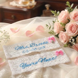 May include: Two white lace garters with blue embroidered text, "Mrs. Brooks 02-28-25" and "You're Next!" are displayed. Each garter has a pink bow. A bouquet of pink roses and scattered petals are also visible.