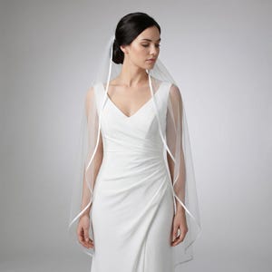 May include: A sheer, white bridal veil with a delicate ribbon trim. The veil is draped over the shoulders and frames the face. The bride is wearing a white, sleeveless wedding dress with a V-neckline. The background is a soft, neutral gray.