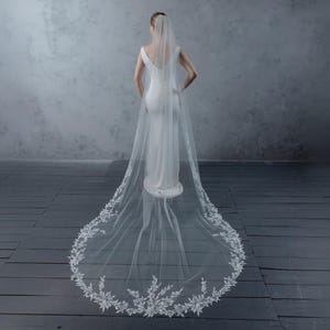 May include: A bride in a white wedding dress and veil, viewed from the back. The veil is long and sheer, with a delicate floral lace trim along the edge. The dress is form-fitting and sleeveless. The background is a neutral gray.