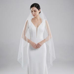 May include: A white wedding dress with a plunging neckline and a sheer, flowing veil. The veil drapes over the shoulders and down the back. The dress is a simple, elegant design, perfect for a wedding ceremony.