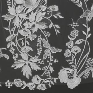 May include: Close-up of white floral lace fabric. The lace features detailed embroidery of flowers, leaves, and butterfly-like designs. Small, sparkling embellishments are scattered throughout the design, adding a touch of elegance. The delicate pattern is set against a dark background.