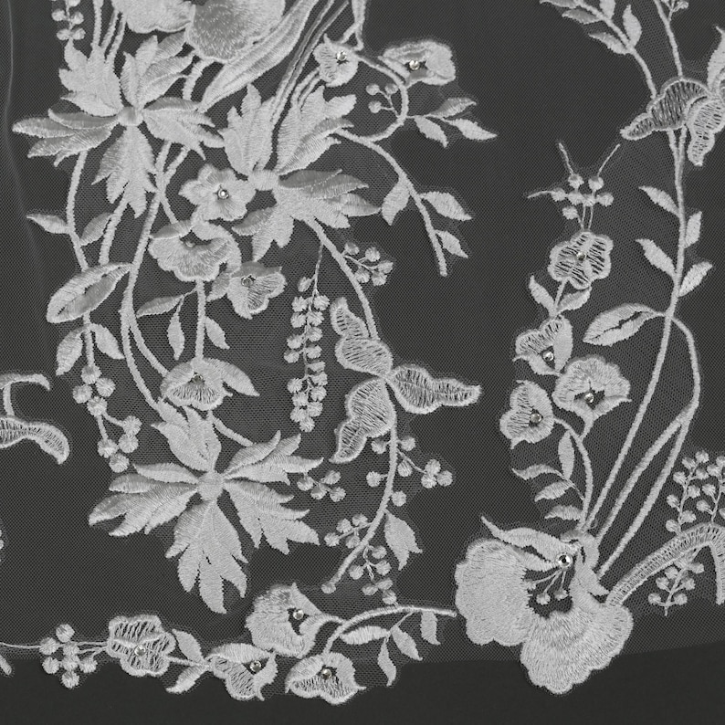 May include: Close-up of white floral lace fabric. The lace features detailed embroidery of flowers, leaves, and butterfly-like designs. Small, sparkling embellishments are scattered throughout the design, adding a touch of elegance. The delicate pattern is set against a dark background.