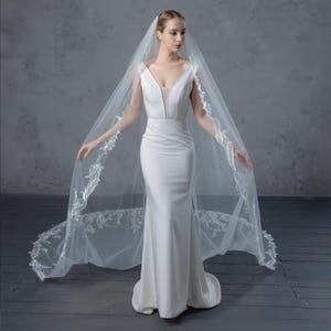 May include: A white wedding dress with a V-neckline and a long, flowing skirt. The bride is wearing a long, sheer veil with delicate floral lace detailing along the edges. The dress is a simple, elegant design.