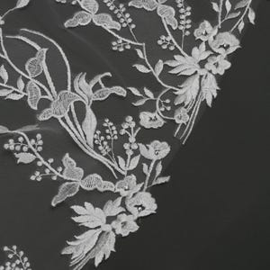 May include: Close-up of white floral lace fabric. The delicate lace features embroidered flowers, leaves, and vines. The design includes various flower types and small, sparkling embellishments. The fabric is set against a dark background, highlighting the intricate details.