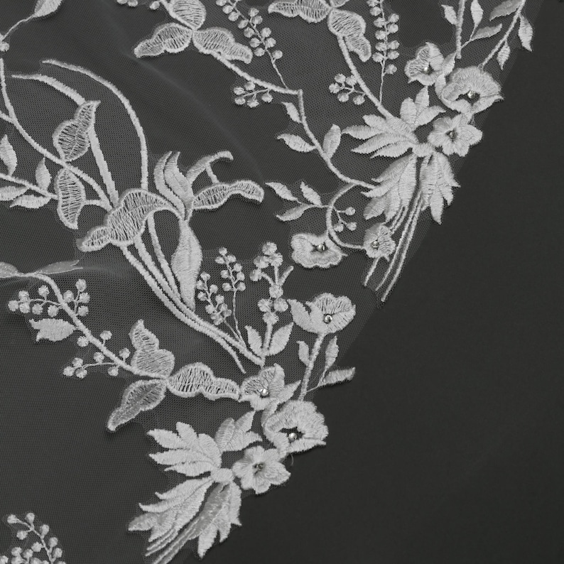 May include: Close-up of white floral lace fabric. The delicate lace features embroidered flowers, leaves, and vines. The design includes various flower types and small, sparkling embellishments. The fabric is set against a dark background, highlighting the intricate details.
