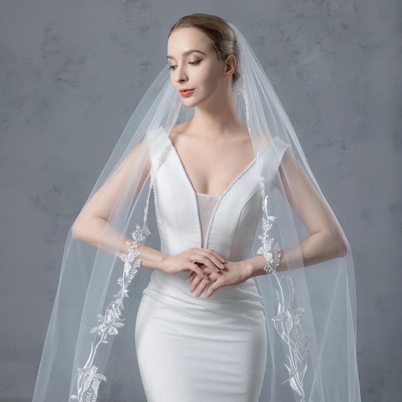 May include: A white wedding dress with a plunging neckline and a long, sheer veil. The veil is adorned with delicate floral embroidery along the edges. The dress is form-fitting and elegant, perfect for a bride.