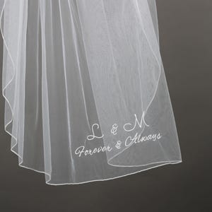 May include: A sheer white wedding veil with delicate embroidery. The veil features the initials "L & M" and the phrase "Forever & Always" in elegant script. The fabric is lightweight and flowing, ideal for a bridal accessory.