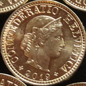 May include: A gold-colored Swiss coin with the inscription "CONFOEDERATIO HELVETICA" and the year "2019" on the front. The coin features a portrait of a woman with a crown.