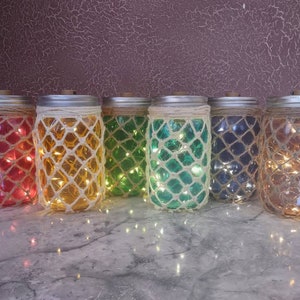 Puede incluir: Seis tarros de vidrio con luces blancas en su interior, cada tarro es de un color diferente: rojo, amarillo, verde, azul, marrón y transparente. Los tarros están sobre una superficie blanca.