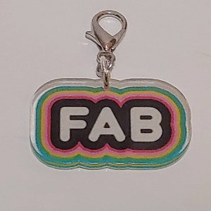 May include: A colorful charm with the word "FAB" in white letters on a black background. The charm has a rainbow-colored border and a silver clasp. The charm is a fun accessory.