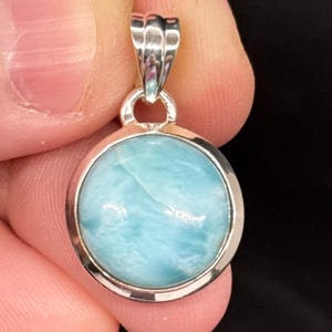May include: A round pendant with a light blue gemstone set in a silver-colored metal. The gemstone has a cloudy appearance. The pendant has a decorative bail for a chain. The background is dark.