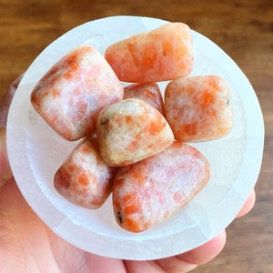 May include: A collection of six sunstone crystals in a white bowl. The crystals are a mix of orange and white, with a translucent appearance.