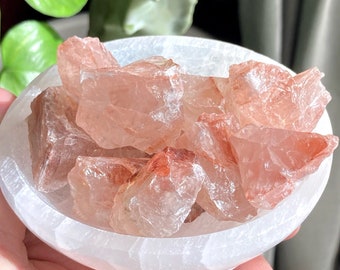 May include: A white selenite bowl filled with a collection of raw, translucent, pink and red crystals. The crystals have a natural, rough texture.