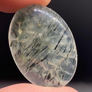 May include: A clear, oval-shaped gemstone with black, needle-like inclusions. The stone is held between two fingers against a dark background. The inclusions are scattered throughout the stone, creating a unique pattern. The gemstone is polished and has a smooth surface.