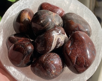 May include: A collection of tumbled red and brown jasper stones with white veins. The stones are smooth and polished, and they are arranged in a white bowl.