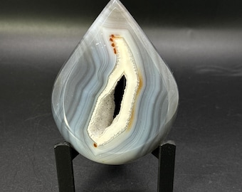 May include: A teardrop-shaped agate stone featuring concentric bands of gray and white, with a central hollow geode cavity lined with small white crystals and accented by thin orange-brown mineral deposits, supported by a black stand.