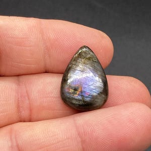 Op de afbeelding: Een traanvormige labradoriet edelsteen, met iriserende flitsen van blauw, paars en goud. De gepolijste steen heeft een glad oppervlak en wordt in een hand gehouden. De edelsteen is ongeveer 2,5 cm lang.