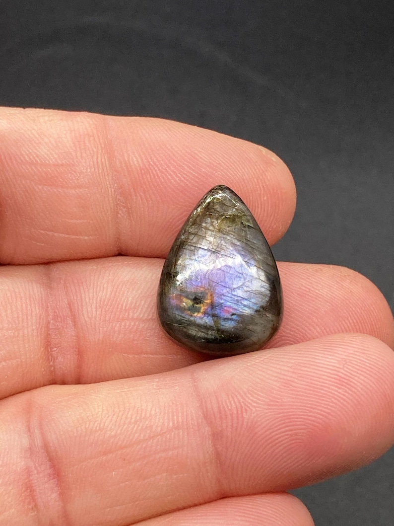 Op de afbeelding: Een traanvormige labradoriet edelsteen, met iriserende flitsen van blauw, paars en goud. De gepolijste steen heeft een glad oppervlak en wordt in een hand gehouden. De edelsteen is ongeveer 2,5 cm lang.