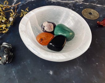 May include: A white selenite bowl with five tumbled stones: green, orange, black, purple, and white. The bowl is on a dark gray surface.