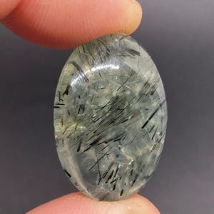 May include: An oval-shaped gemstone with a translucent, pale green base and striking black needle-like inclusions. The polished surface reflects light, highlighting the internal patterns. The gemstone is held between two fingers.
