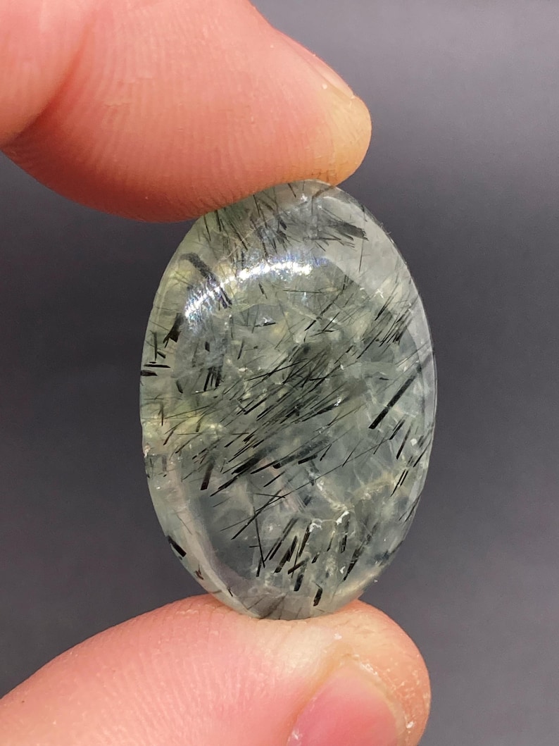 May include: An oval-shaped gemstone with a translucent, pale green base and striking black needle-like inclusions. The polished surface reflects light, highlighting the internal patterns. The gemstone is held between two fingers.