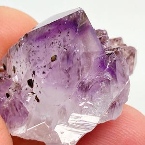 May include: A cluster of amethyst crystals with a purple hue and black inclusions. The crystals are pointed and have a natural, rough texture.
