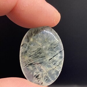 May include: An oval-shaped gemstone with a translucent, pale green base. The stone is filled with black, needle-like inclusions, creating a striking contrast. The gemstone is held between two fingers against a dark background.