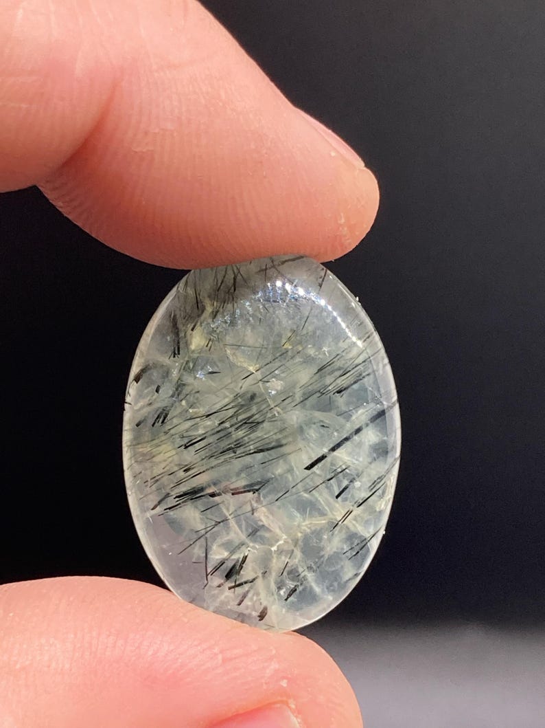 May include: An oval-shaped gemstone with a translucent, pale green base. The stone is filled with black, needle-like inclusions, creating a striking contrast. The gemstone is held between two fingers against a dark background.