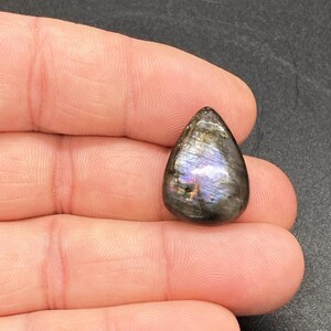 Op de afbeelding: Een traanvormige labradoriet edelsteen, gepolijst en met iriserende flitsen van blauw, paars en goud. De steen wordt in een hand gehouden, waardoor de natuurlijke schoonheid en het unieke kleurenspel worden benadrukt. Een natuurlijke, gepolijste steen.