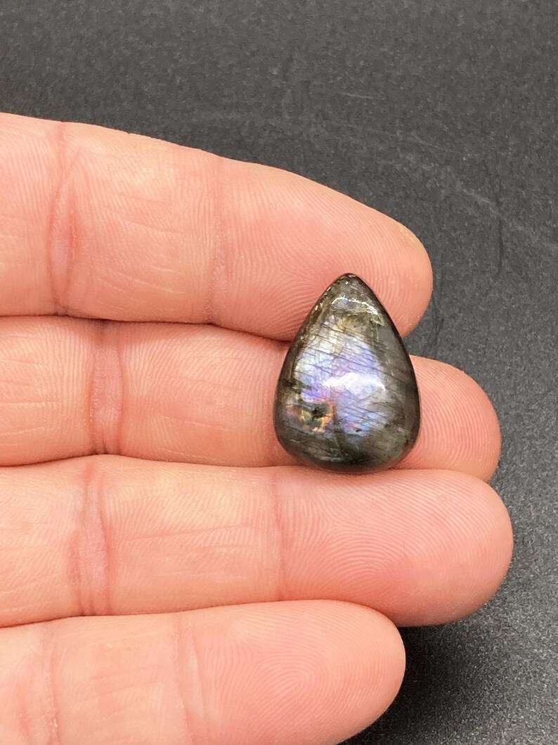 Op de afbeelding: Een traanvormige labradoriet edelsteen, gepolijst en met iriserende flitsen van blauw, paars en goud. De steen wordt in een hand gehouden, waardoor de natuurlijke schoonheid en het unieke kleurenspel worden benadrukt. Een natuurlijke, gepolijste steen.