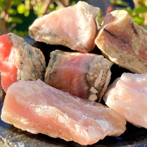 May include: A collection of pink and white rough opal stones. The stones are various sizes and shapes, with some showing a more opaque white color and others a more translucent pink color.