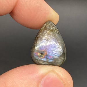 Op de afbeelding: Een traanvormige labradoriet edelsteen, vastgehouden tussen vingers, toont iriserende flitsen van blauw, paars en goud. De gepolijste steen heeft een glad oppervlak en een grijze basiskleur, met een subtiele glans. De edelsteen is een natuurlijk mineraal.