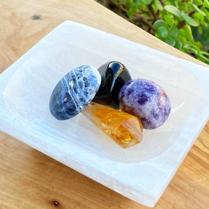May include: A collection of polished gemstones, including a blue sodalite, black obsidian, purple lepidolite, and a golden citrine crystal, displayed in a white selenite bowl. The bowl sits on a wooden surface.