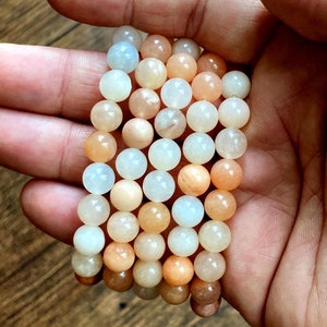 May include: A stack of four bracelets made with round, smooth, sunstone beads in shades of white, peach, and orange.