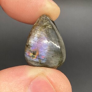 Op de afbeelding: Een traanvormige labradoriet edelsteen, vastgehouden tussen vingers. De gepolijste steen toont iriserende flitsen van paars, blauw en goud in een grijze en bruine matrix. De edelsteen is glad en reflecterend en toont natuurlijke patronen.