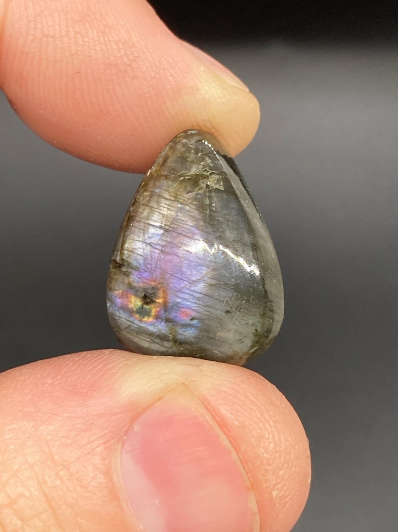 Op de afbeelding: Een traanvormige labradoriet edelsteen, vastgehouden tussen vingers. De gepolijste steen toont iriserende flitsen van paars, blauw en goud in een grijze en bruine matrix. De edelsteen is glad en reflecterend en toont natuurlijke patronen.