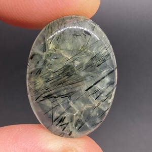 May include: Oval-shaped gemstone with a translucent, pale green base and striking black needle-like inclusions. The polished surface reflects light, showcasing the internal patterns. The gemstone is held between two fingers.