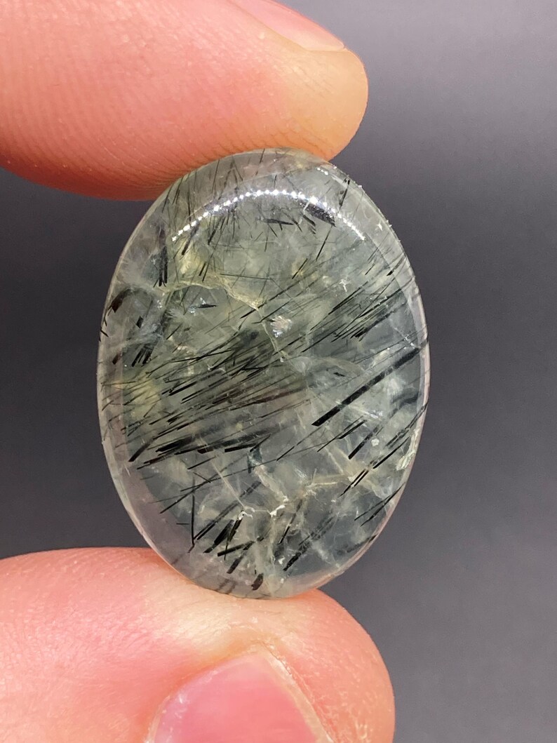 May include: Oval-shaped gemstone with a translucent, pale green base and striking black needle-like inclusions. The polished surface reflects light, showcasing the internal patterns. The gemstone is held between two fingers.