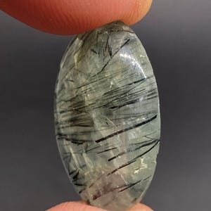 May include: An oval-shaped gemstone with a translucent, pale green base and striking black needle-like inclusions. The stone is held between two fingers, showcasing its polished surface and natural patterns. The gemstone is a unique piece of jewelry.
