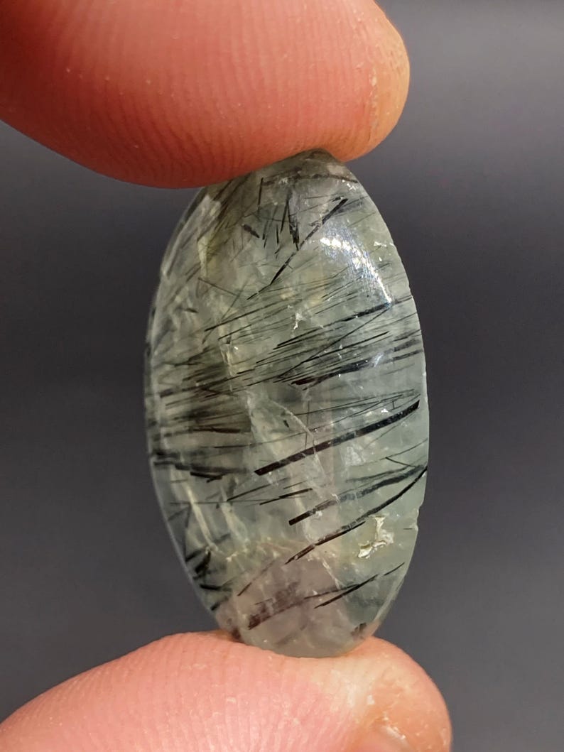 May include: An oval-shaped gemstone with a translucent, pale green base and striking black needle-like inclusions. The stone is held between two fingers, showcasing its polished surface and natural patterns. The gemstone is a unique piece of jewelry.