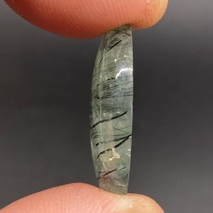 May include: A translucent, oval-shaped gemstone with internal black inclusions. The stone is held between two fingers against a dark gray background. The gemstone has a polished surface and a slightly curved shape.