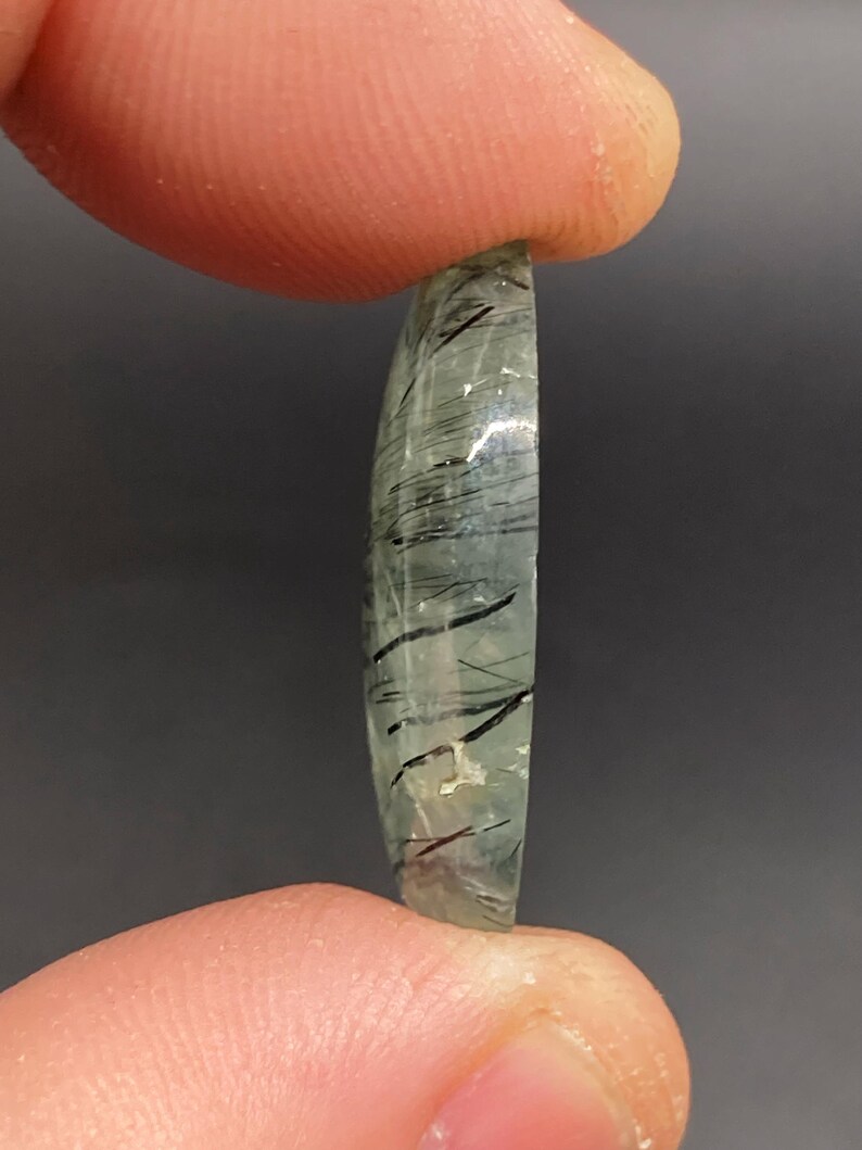 May include: A translucent, oval-shaped gemstone with internal black inclusions. The stone is held between two fingers against a dark gray background. The gemstone has a polished surface and a slightly curved shape.