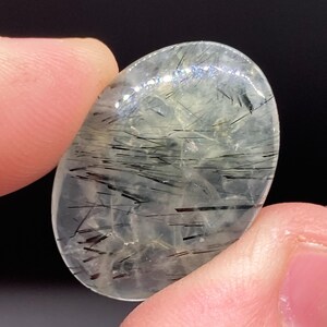 May include: A clear, oval-shaped gemstone with internal black needle-like inclusions. The stone is held between two fingers against a dark background. The inclusions create a striking contrast within the translucent material.
