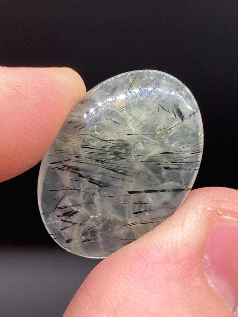 May include: A clear, oval-shaped gemstone with internal black needle-like inclusions. The stone is held between two fingers against a dark background. The inclusions create a striking contrast within the translucent material.